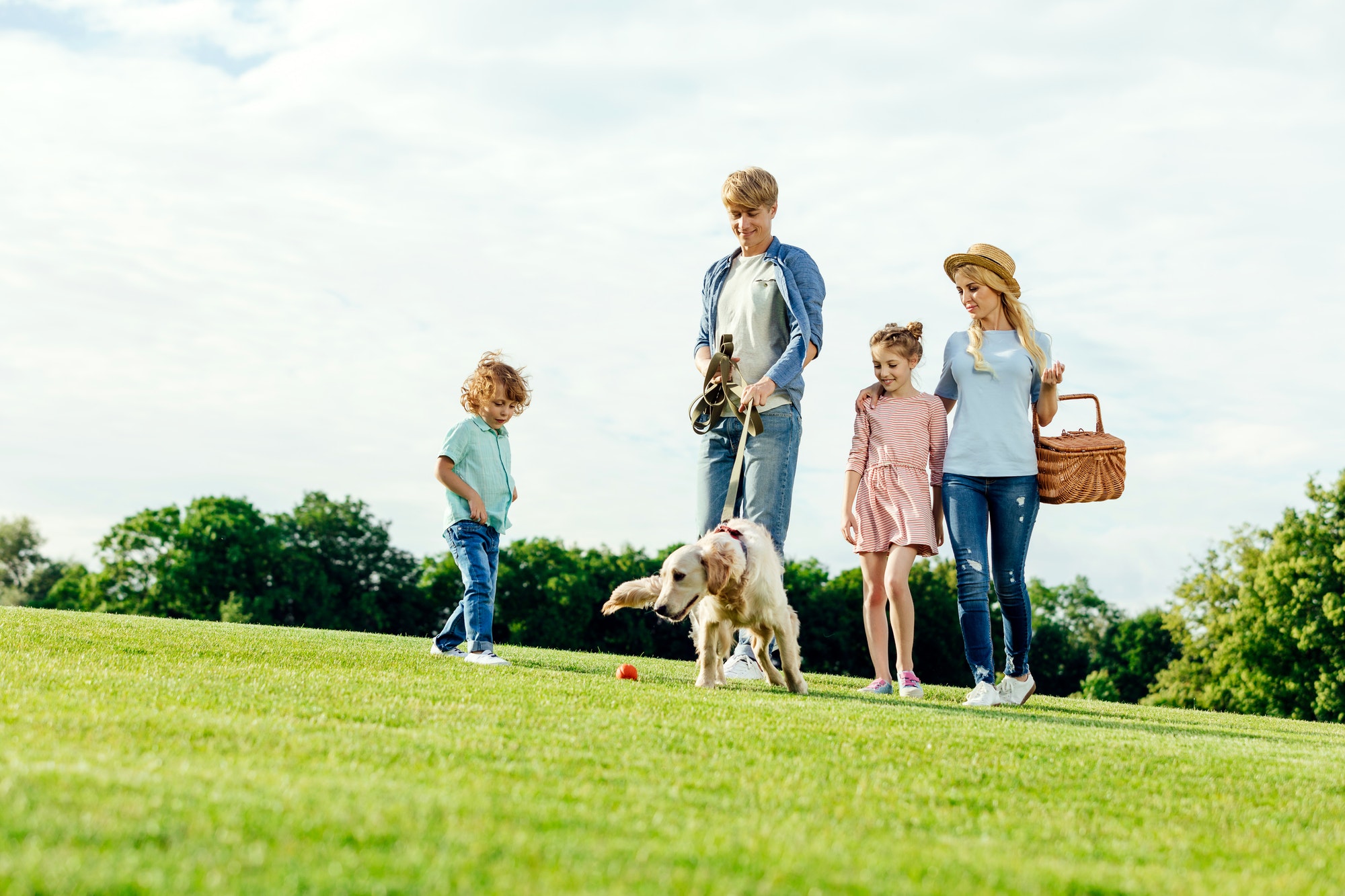 happy young family with picnic basket walking with dog on green lawn